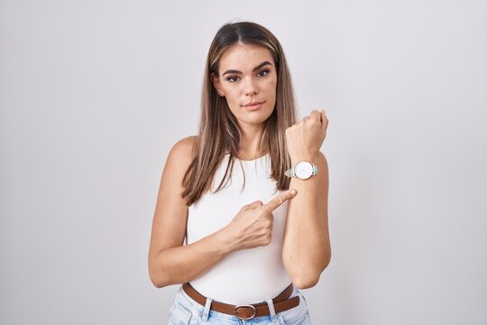 Hispanic young woman standing over white background in hurry pointing to watch time, impatience, looking at the camera with relaxed expression