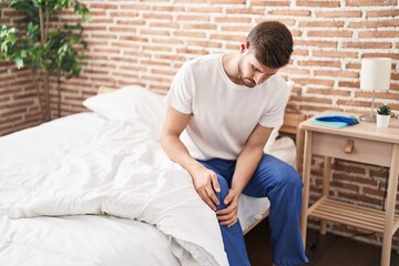 Young caucasian man suffering for knee injury sitting on bed at bedroom