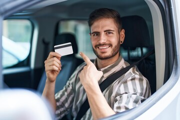 Hispanic man with beard driving car holding credit card smiling happy pointing with hand and finger