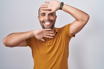 Hispanic man with beard standing over white background smiling cheerful playing peek a boo with...