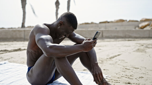 African american man tourist sitting on towel shirtless using smartphone at the beach - Powered by Adobe