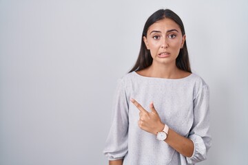Young hispanic woman standing over white background pointing aside worried and nervous with forefinger, concerned and surprised expression - obrazy, fototapety, plakaty