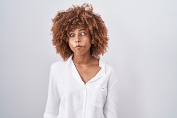 Young hispanic woman with curly hair standing over white background making fish face with lips, crazy and comical gesture. funny expression.