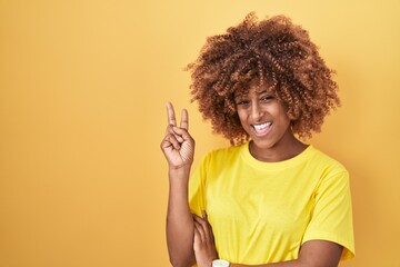Young hispanic woman with curly hair standing over yellow background smiling with happy face winking at the camera doing victory sign. number two. - obrazy, fototapety, plakaty