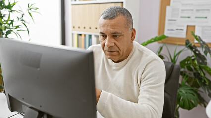 A mature hispanic man focusing on work in a modern office setting with plants and a computer...