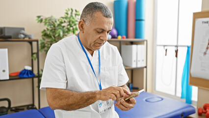 A middle-aged hispanic man in a white coat uses a smartphone in a brightly-lit rehabilitation...