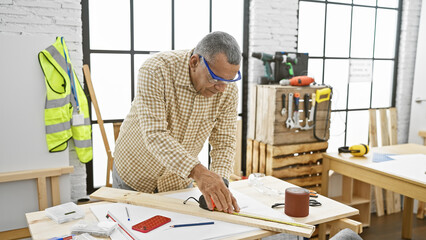 A mature hispanic man measuring wood in a bright carpentry workshop.