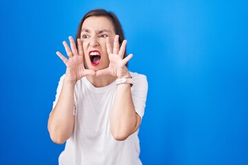Middle age hispanic woman standing over blue background shouting angry out loud with hands over mouth