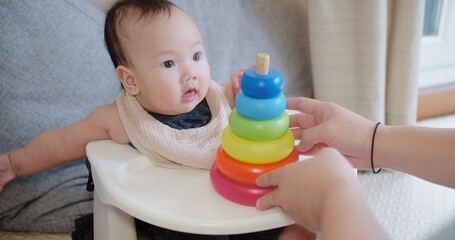 Joyful infant baby sits in a high chair learning to stack colorful rings with help from mother, a moment engaging in early childhood development activities.