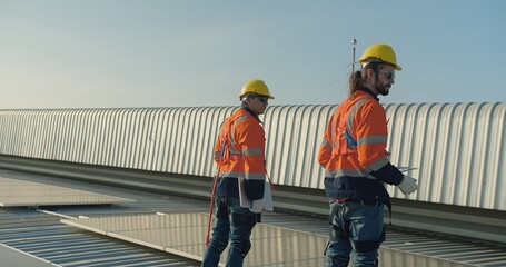 Two solar engineers in reflective gear discuss work plans project details on a rooftop with solar...