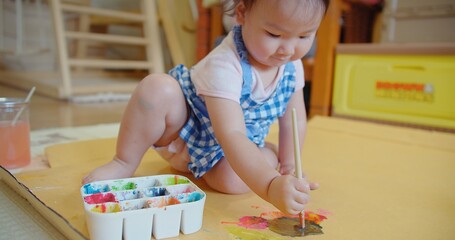 An engrossed toddler in a blue checkered dress discovers creativity with a paintbrush in hand and...
