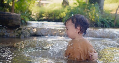 A curious toddler in a brown shirt joyfully explores the sensations of splashing water experiences the joy of water play in a shallow stream, surrounded by lush vegetation