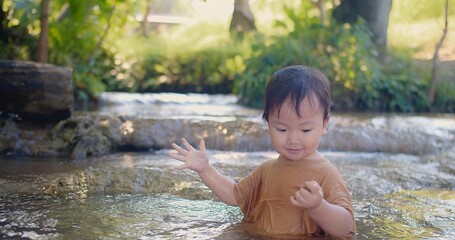 A curious toddler in a brown shirt joyfully explores the sensations of splashing water experiences the joy of water play in a shallow stream, surrounded by lush vegetation