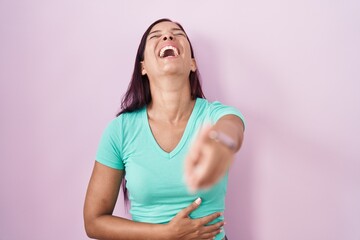Young hispanic woman standing over pink background laughing at you, pointing finger to the camera...