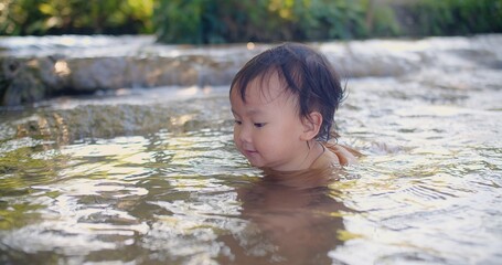 A curious toddler in a brown shirt joyfully explores the sensations of splashing water experiences the joy of water play in a shallow stream, surrounded by lush vegetation