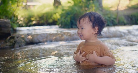 A curious toddler in a brown shirt joyfully explores the sensations of splashing water experiences the joy of water play in a shallow stream, surrounded by lush vegetation