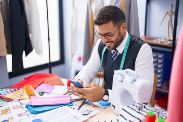 Young hispanic man tailor smiling confident using smartphone at tailor shop