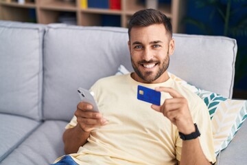 Young hispanic man using smartphone and credit card sitting on sofa at home