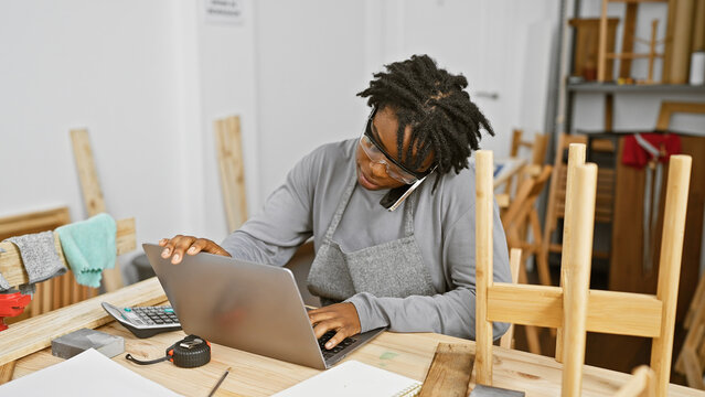 Young Black Woman With Dreadlocks Multitasking In A Carpentry Workshop, Using A Laptop While Talking On The Phone.