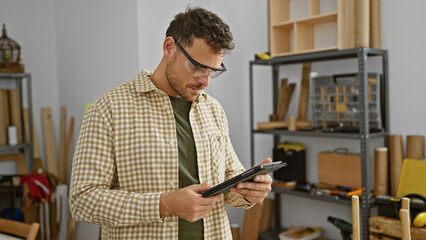 A focused young man with a beard checks a tablet in a well-equipped carpentry workshop.