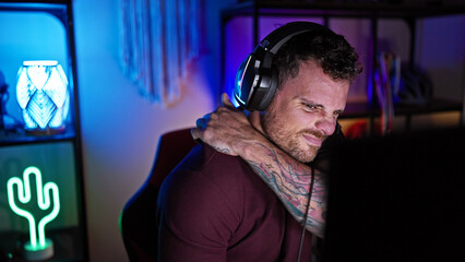 Hispanic man with beard feeling back pain in a dark gaming room at night, wearing headphones.
