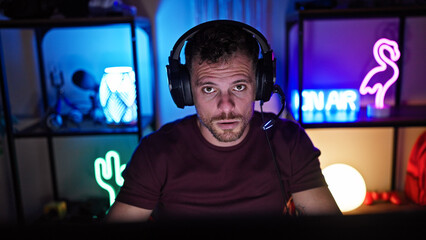 A handsome hispanic man with headphones in a dark gaming room looks intently at the camera
