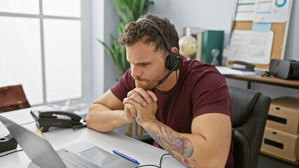 A focused hispanic man with a beard and tattoo works indoors at an office wearing a headset in front of a laptop.