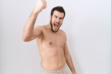 Young hispanic man standing shirtless over white background angry and mad raising fist frustrated and furious while shouting with anger. rage and aggressive concept.