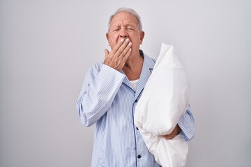Senior man with grey hair wearing pijama hugging pillow bored yawning tired covering mouth with...