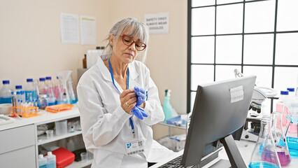 Mature woman scientist in lab coat analyzing data on computer amidst laboratory equipment.