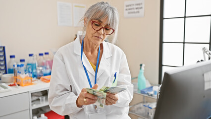 Mature woman in white lab coat counting hong kong dollars at a laboratory indoors.
