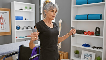Mature woman exercises with dumbbells in a rehab clinic's interior, showcasing healthcare, fitness, and well-being for middle-aged adults.