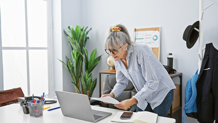 Mature woman with grey hair working in a modern office, standing at her desk with papers and a...