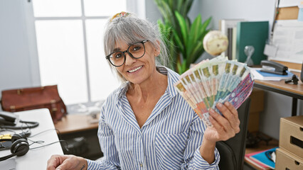 Smiling mature woman displaying hong kong dollars in a modern office environment.