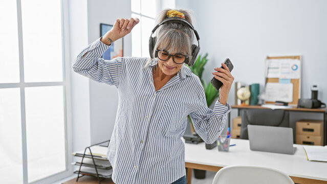 A joyful middle-aged woman with grey hair dances in an office room, wearing headphones and holding a smartphone.