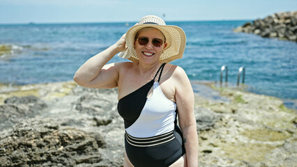 Middle age grey-haired woman tourist wearing swimsuit and summer hat smiling at the beach