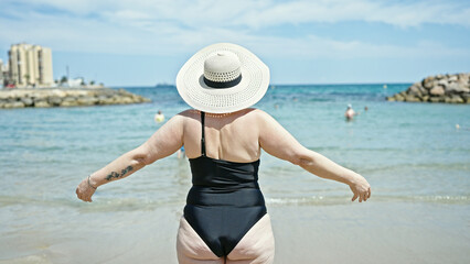 Middle age grey-haired woman tourist wearing swimsuit and summer hat standing with arms open backwards at the beach