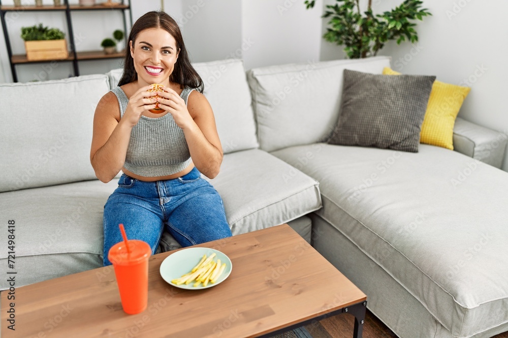 Poster Young beautiful hispanic woman eating hamburger sitting on sofa at home