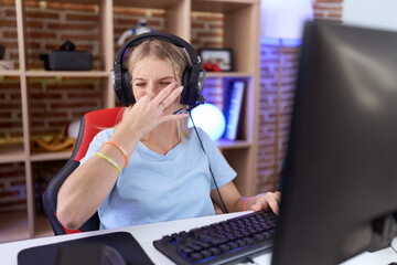Young caucasian woman playing video games wearing headphones smelling something stinky and...