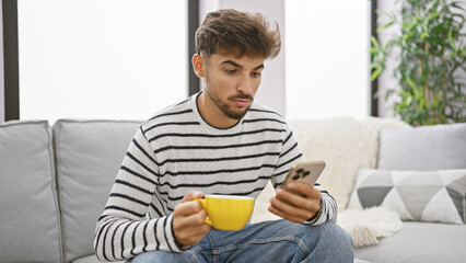 Handsome young arab man relaxing at home, sipping coffee and using smartphone, displaying a serious expression as he immerses in online communication, comfortably resting on his living room sofa. - obrazy, fototapety, plakaty
