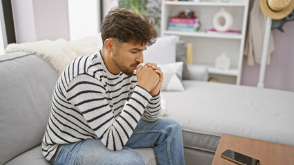 Worried young arab man with a beard, sitting on his living room sofa at home, seriously expressing...