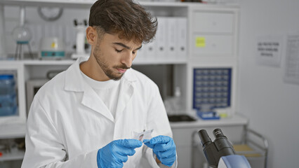 Arabian young man scientist, deeply concentrated in his analysis, looking at a sample in lab with focus, balancing research and safety with professionalism in science - obrazy, fototapety, plakaty