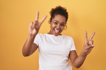 Young hispanic woman with curly hair standing over yellow background smiling with tongue out...
