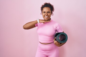 Young hispanic woman with curly hair holding yoga mat over pink background cutting throat with hand...