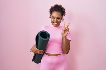 Young hispanic woman with curly hair holding yoga mat over pink background smiling with happy face...