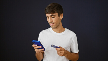 Casually-dressed handsome hispanic man standing against isolated black background, totally immersed, using smartphone and credit card for online shopping
