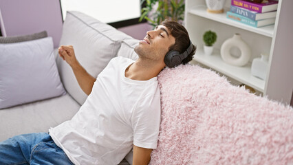 Portrait of a relaxed, confident young hispanic man, joyfully enjoying the sound of music, listening through headphones, sitting indoors on the sofa in a cosy, modern apartment.