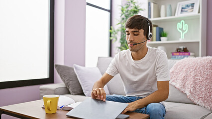 At home, young handsome hispanic man, a dedicated call center agent, opening laptop, gearing up with headphones, setting the stage in his living room to ace his support role.