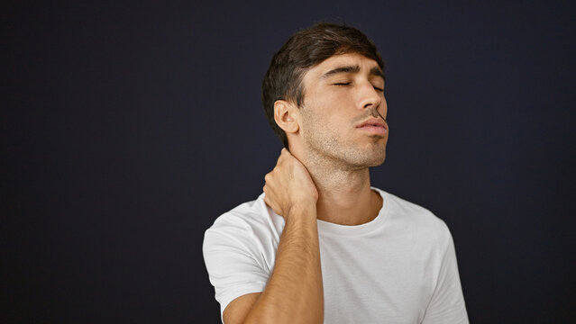 Handsome Young Hispanic Guy Suffering Serious Backache, Standing In Relaxed Concentration, Tired And Stressed Touching Injured Neck On Isolated Black Background, Portrait Looking Away From Camera