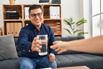 Young latin man psychologist giving glass of water to patient at psychology clinic